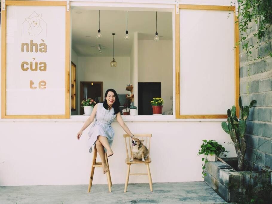 Người phụ nữ ngồi cạnh chú chó trên hai ghế gỗ, trước ô cửa màu hồng có chậu cây và xương rồng