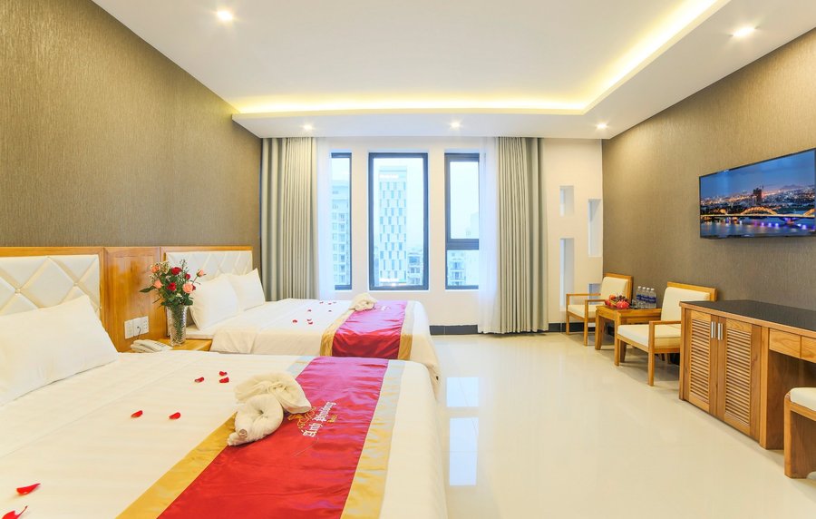 Phòng ngủ với hai giường đôi, khăn trải đỏ, cửa sổ lớn nhìn ra các tòa nhà và TV treo tường