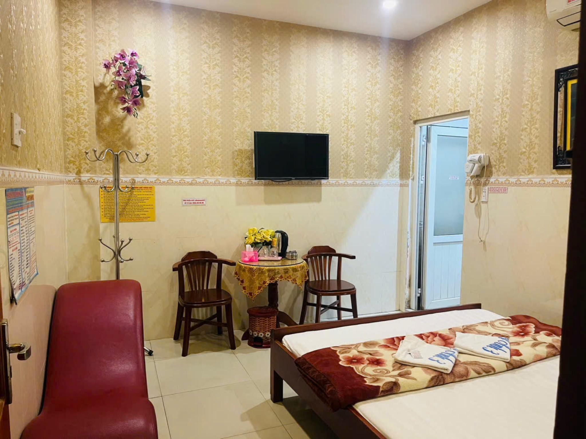 Phòng nghỉ có giường trải chăn hoa, bàn tròn phủ khăn vàng, ghế gỗ và tivi treo tường