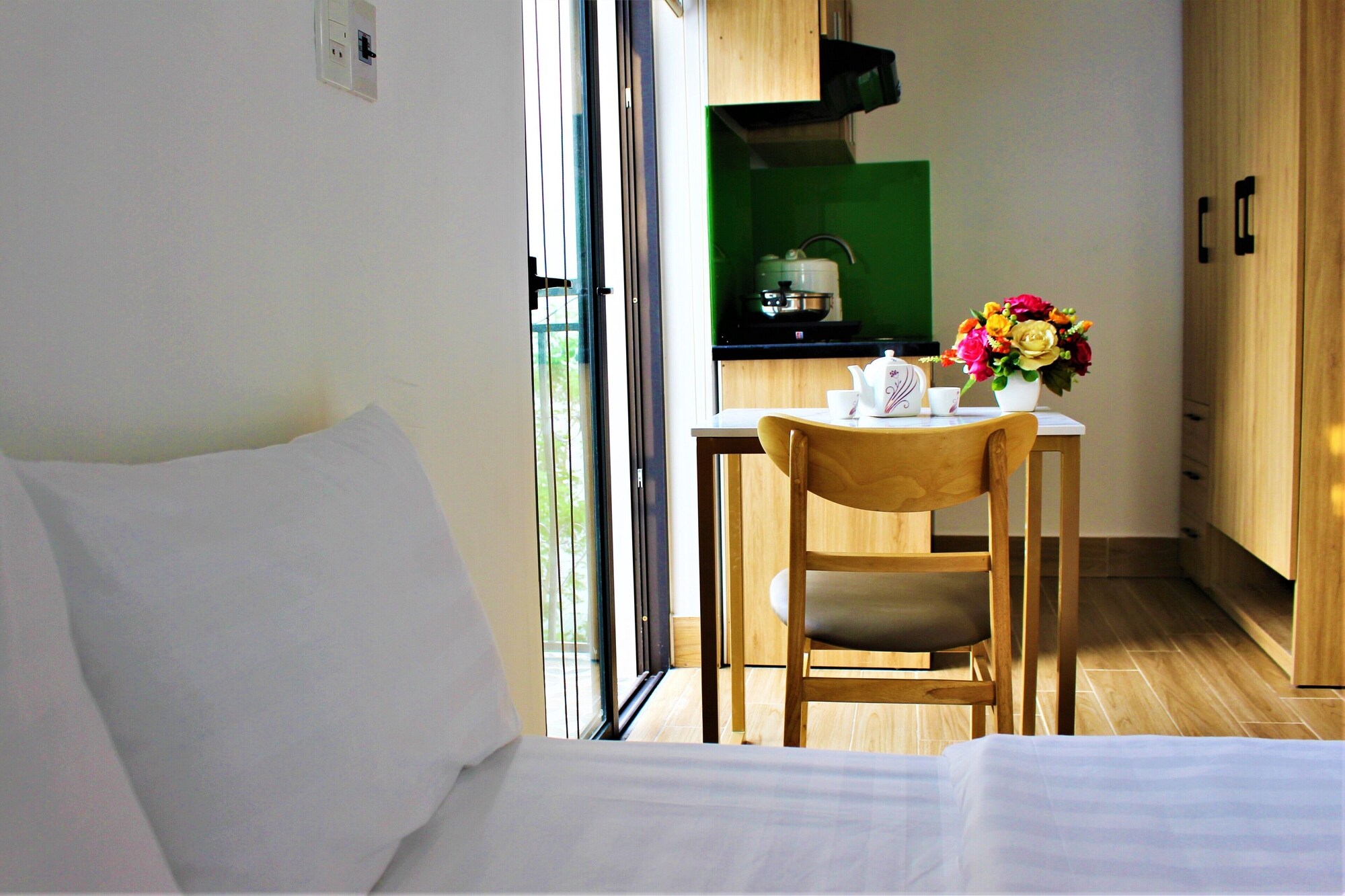 Phòng căn hộ với giường ga trắng, bàn gỗ đặt bộ ấm trà và bình hoa, phía sau là bếp nhỏ màu xanh