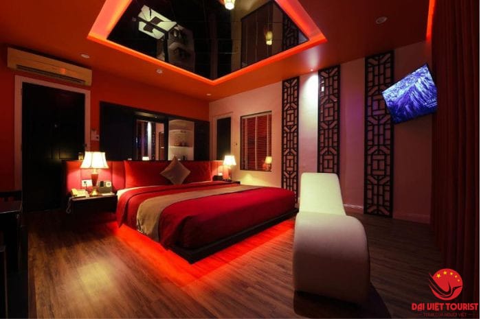 Phòng ngủ giường đôi với ga đỏ, đèn vàng ấm, ghế trắng và tivi treo trên tường