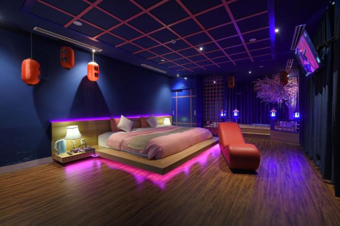 Phòng ngủ tông xanh tím với giường đôi đặt trên bục gỗ, đèn LED hồng và ghế bọc da đỏ