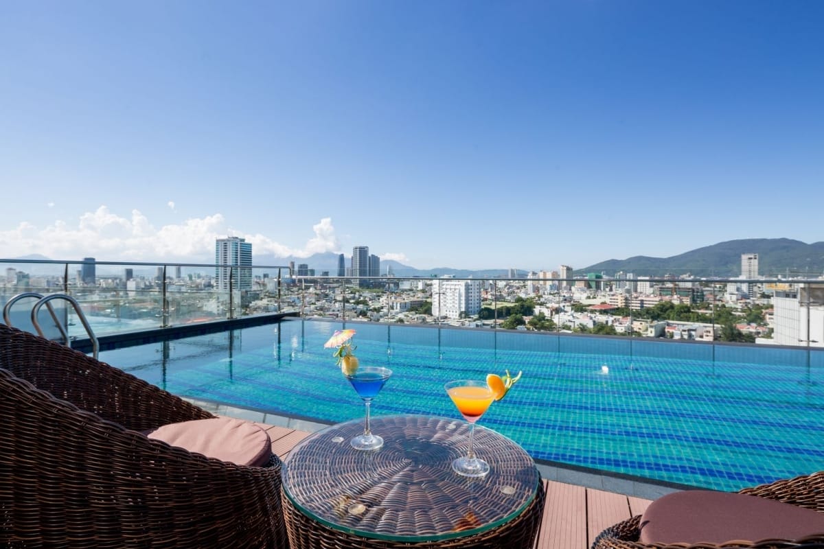 Hồ bơi vô cực trên sân thượng nhìn ra thành phố và núi, cạnh bàn mây có hai ly cocktail