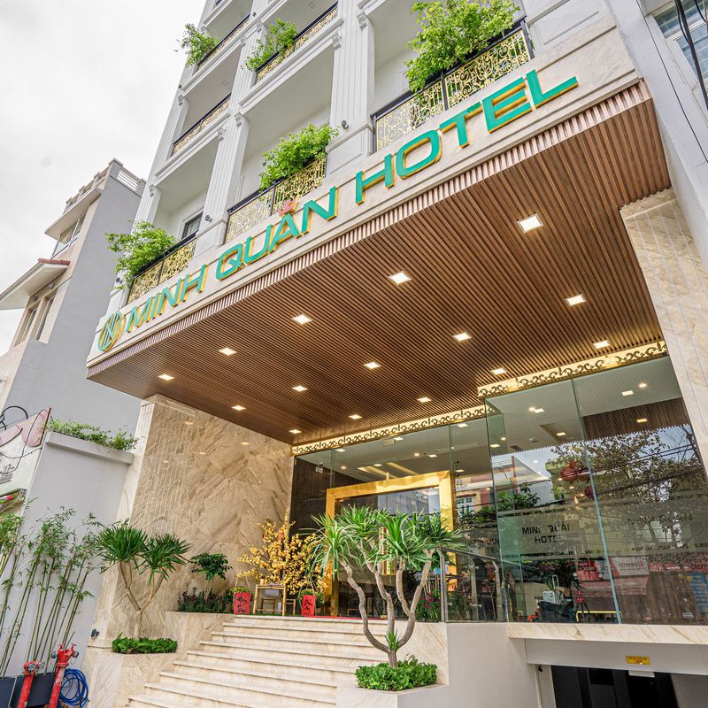 Mặt tiền Minh Quan Hotel nhìn từ dưới lên, biển tên xanh vàng, ban công trắng phủ cây và sảnh kính