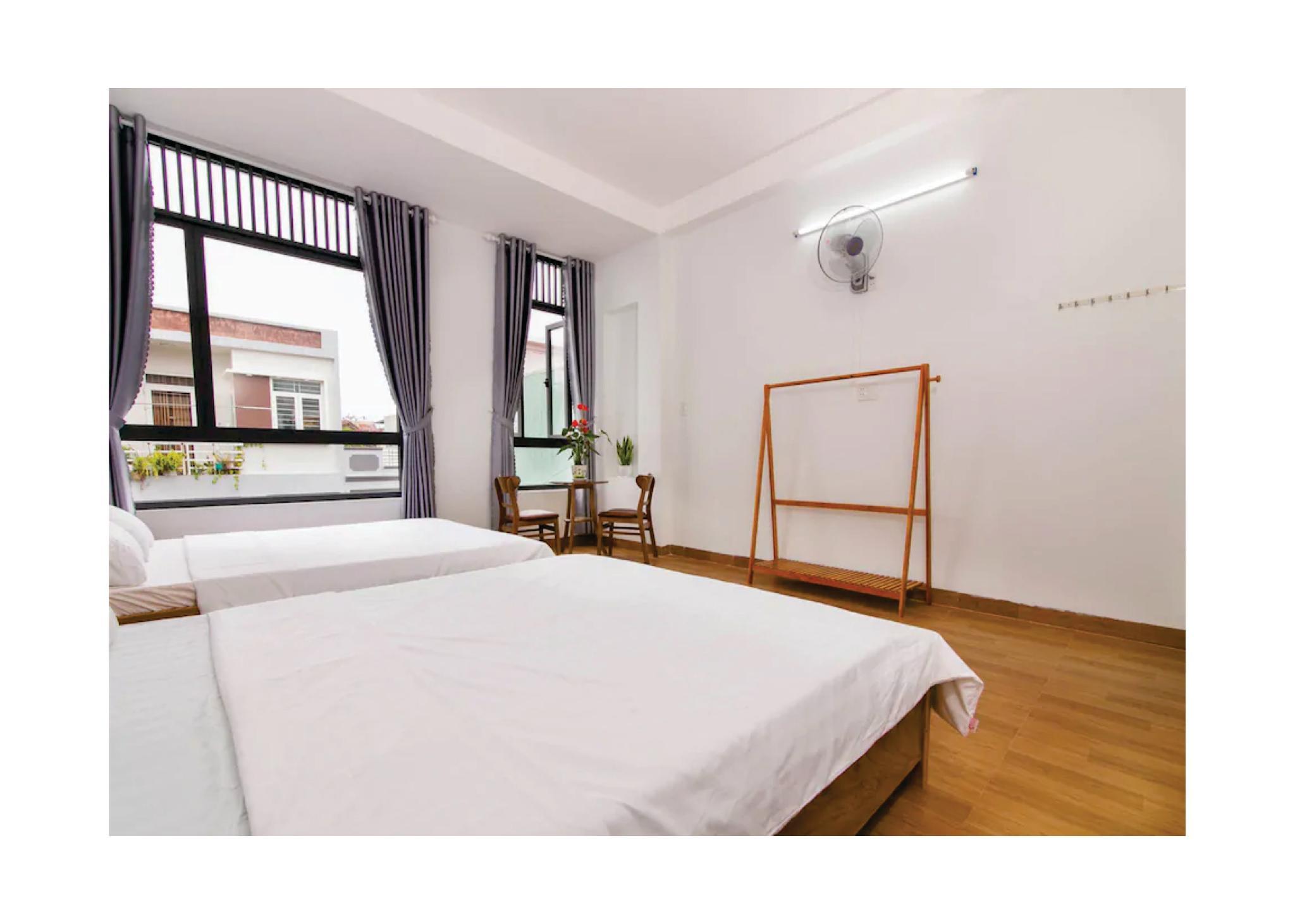 Phòng ngủ sáng với ba giường trải ga trắng, hai cửa sổ rèm xám, bàn ghế gỗ và giá treo đồ