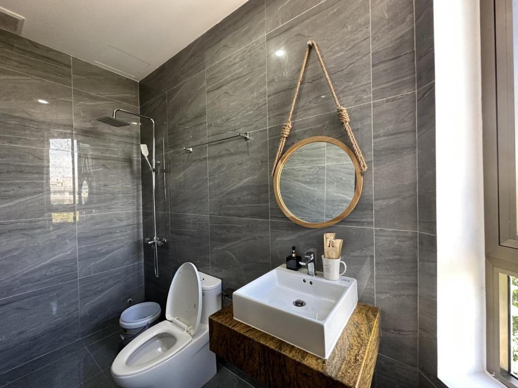 Phòng tắm lát gạch vân đá xám, gồm bồn rửa trắng, bồn cầu, vòi sen và gương tròn treo dây thừng